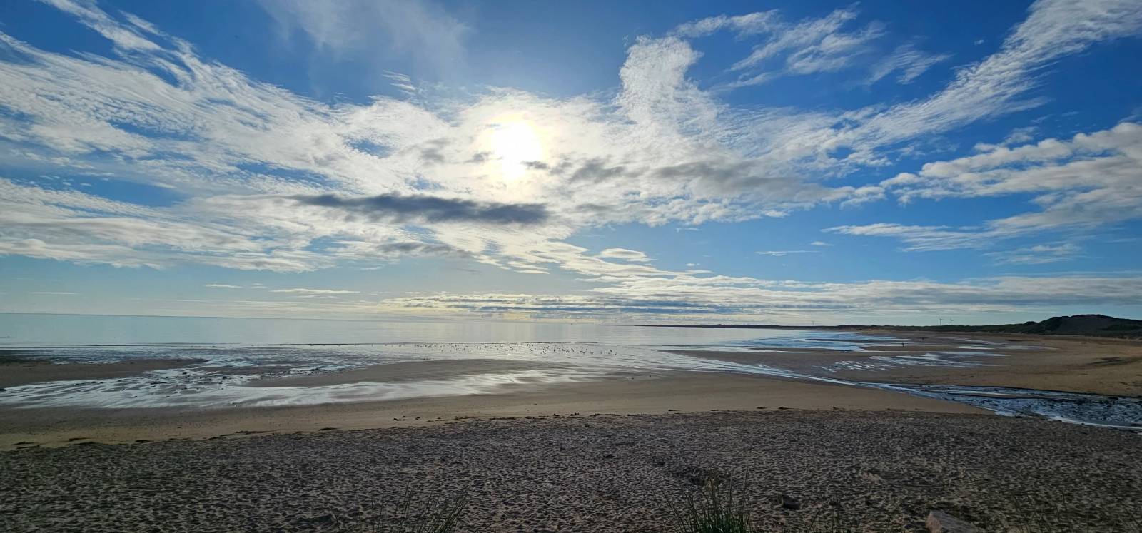 An Aberdeenshire beach, with blue sky, sun and clouds reflecting on the calm sea below.